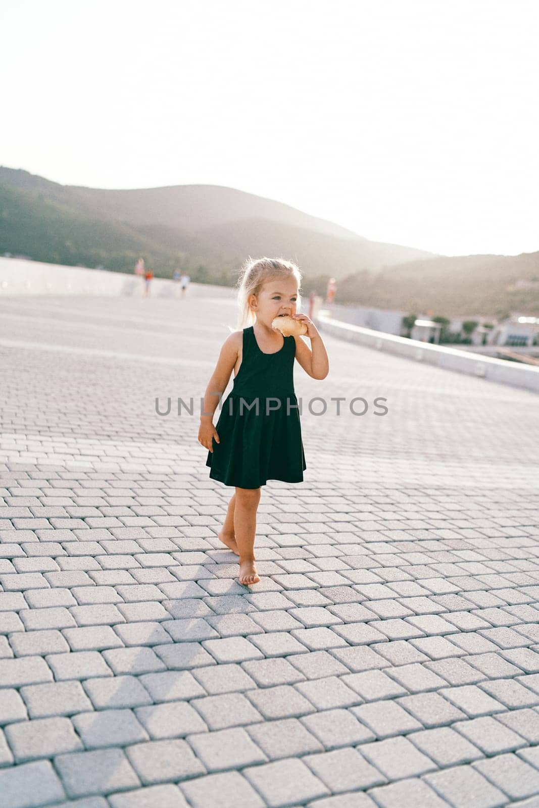 Little girl nibbles on a bun while walking barefoot on a tiled road. High quality photo