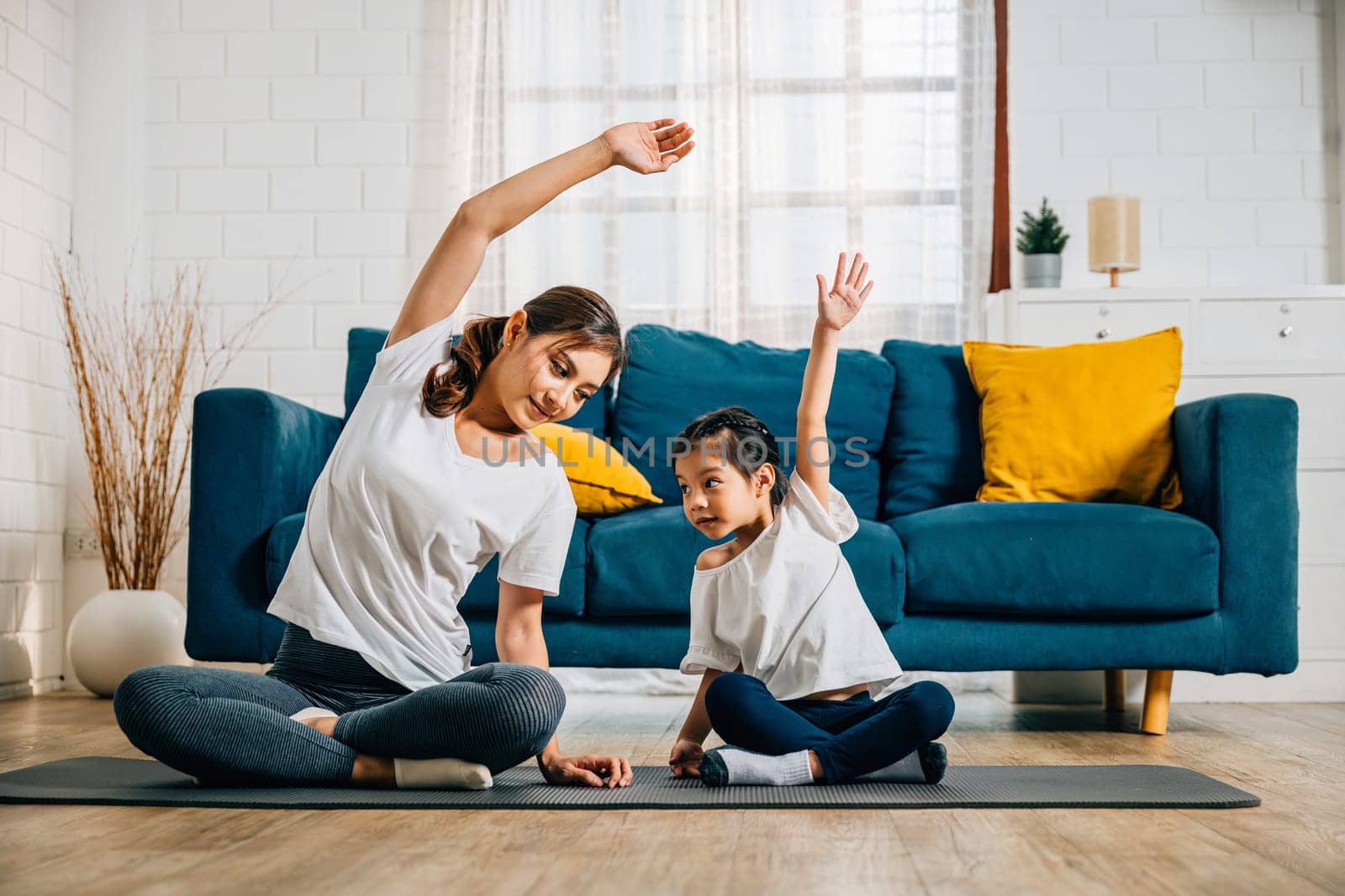 A mother supports her little daughter's yoga practice creating a happy family moment of relaxation bonding and education. Their smiles and concentration highlight domestic togetherness and wellness.