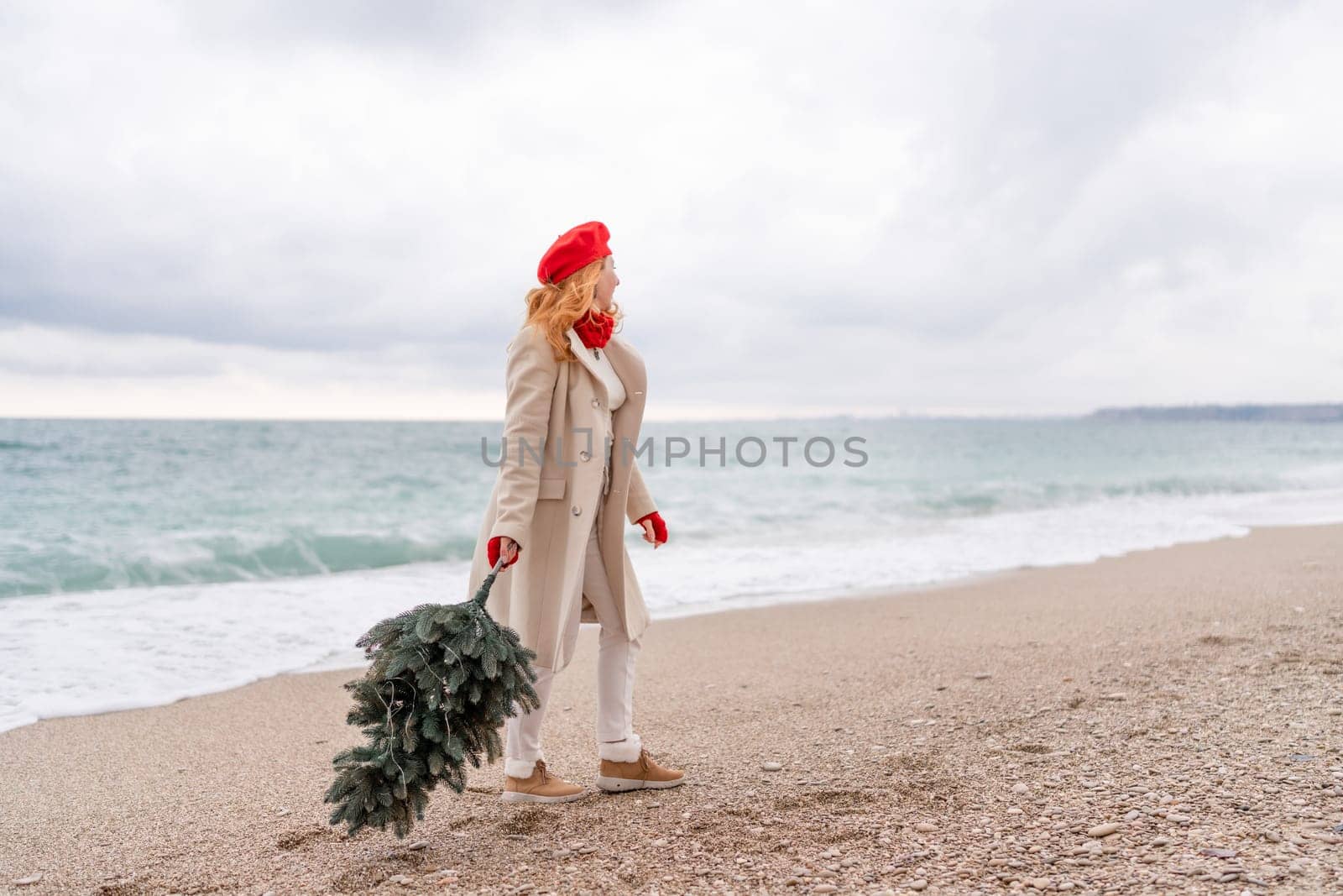 Redhead woman Christmas tree sea. Christmas portrait of a happy redhead woman walking along the beach and holding a Christmas tree in her hands. She is dressed in a light coat and a red beret