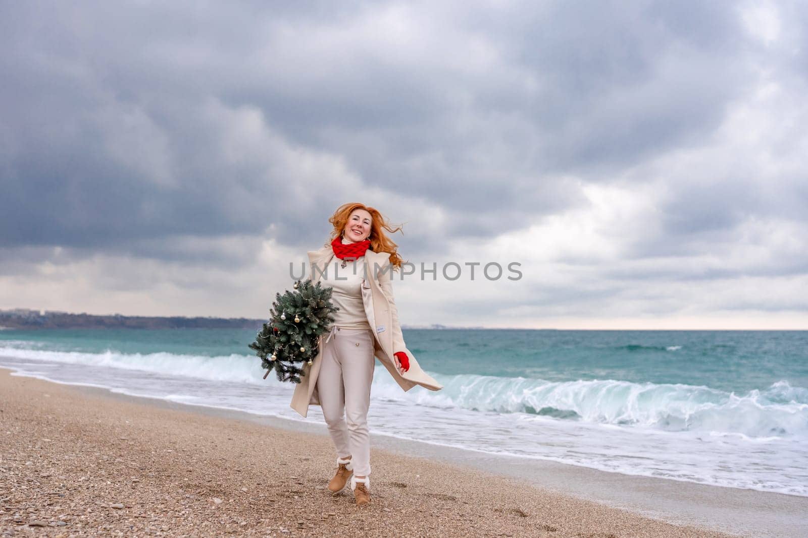 Redhead woman Christmas tree sea. Christmas portrait of a happy redhead woman walking along the beach and holding a Christmas tree in her hands. Dressed in a light coat, white suit and red mittens