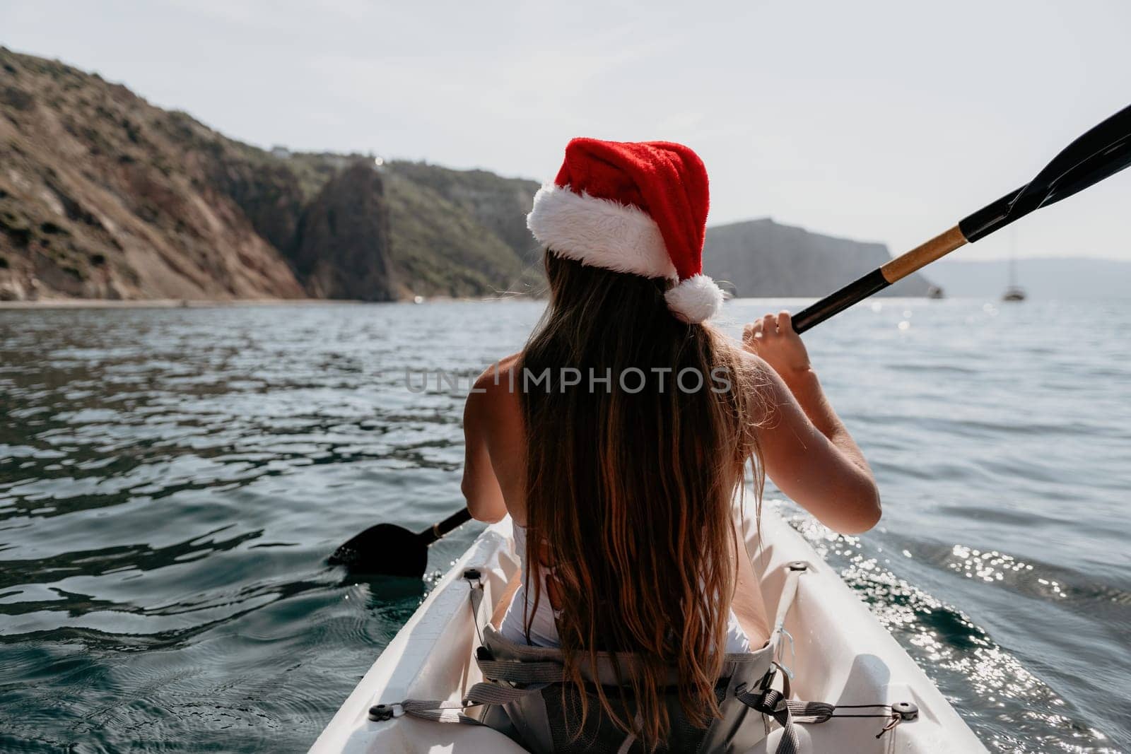 Woman in kayak back view. Happy young woman in Santa hat floating in kayak on calm sea. Summer holiday vacation and cheerful female people relaxing having fun on the boat. by panophotograph