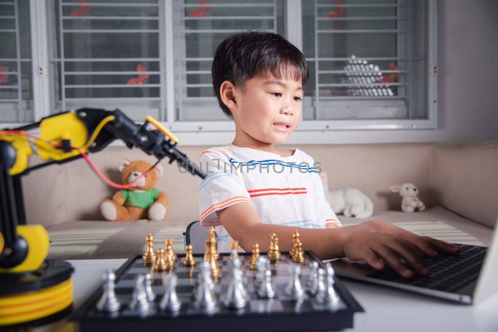 Asian little boy programming code to robot machine arm on laptop for play chess by Sorapop