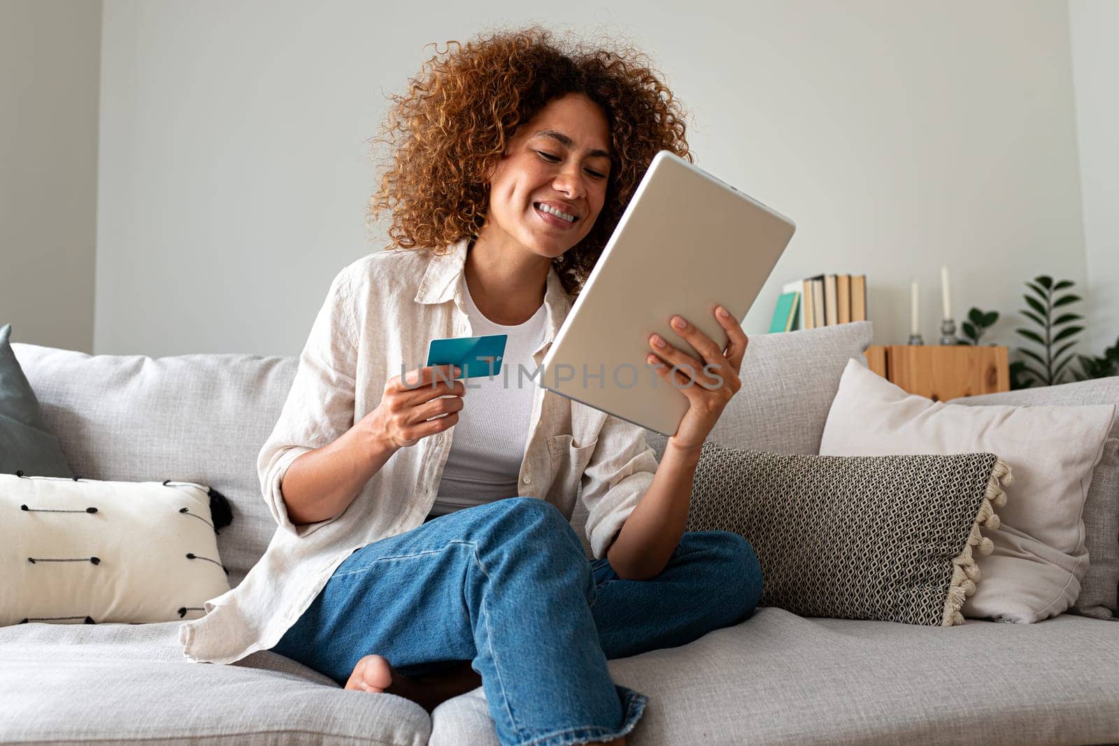 Happy and smiling young Multiracial hispanic woman online shopping using digital tablet and credit card sitting on the couch at home. E-commerce concept.