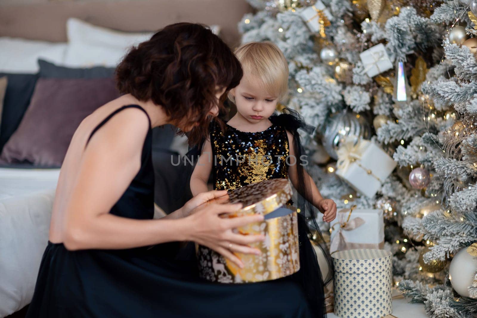Mother daughter 2 years old Christmas tree. Both are dressed in black dresses, the mother holds the girl in her arms and both look at each other. The family celebrates Christmas. by Matiunina