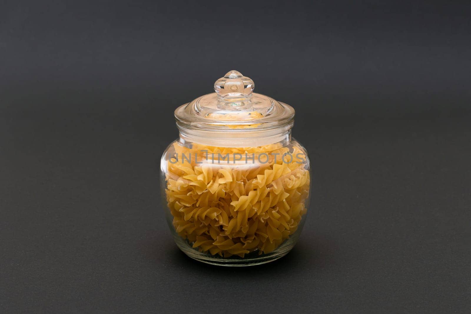 Uncooked Fusilli Pasta in Glass Jar on Black Background. Raw and Dry Macaroni. Unhealthy and Fat Food. Italian Culture