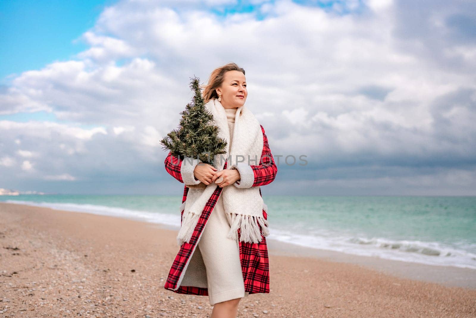 Blond woman holding Christmas tree by the sea. Christmas portrait of a happy woman walking along the beach and holding a Christmas tree in her hands. Dressed in a red coat, white dress