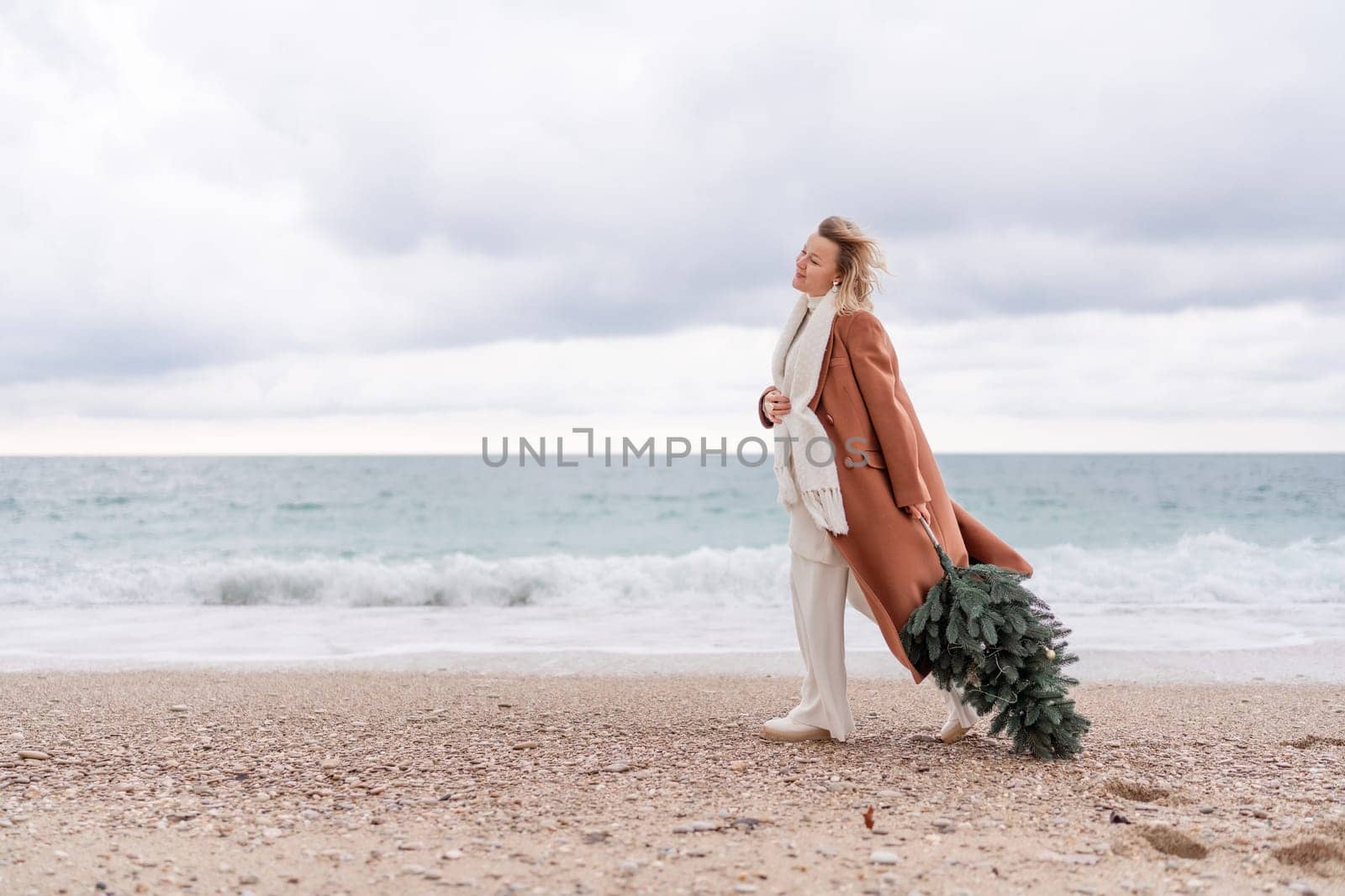 Blond woman Christmas sea. Christmas portrait of a happy woman walking along the beach and holding a Christmas tree in her hands. She is wearing a brown coat and a white suit