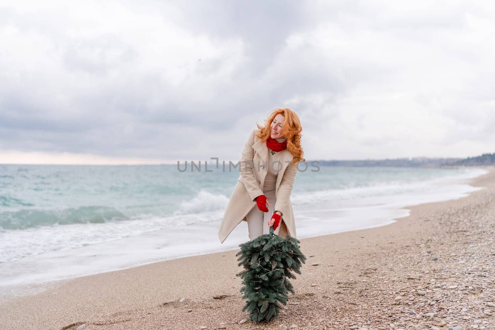 Redhead woman Christmas tree sea. Christmas portrait of a happy redhead woman walking along the beach and holding a Christmas tree in her hands. Dressed in a light coat, white suit and red mittens. by Matiunina