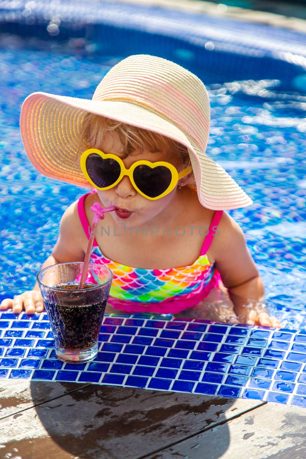 A child near the pool in sunglasses drinks a cocktail. Selective focus. Kid.