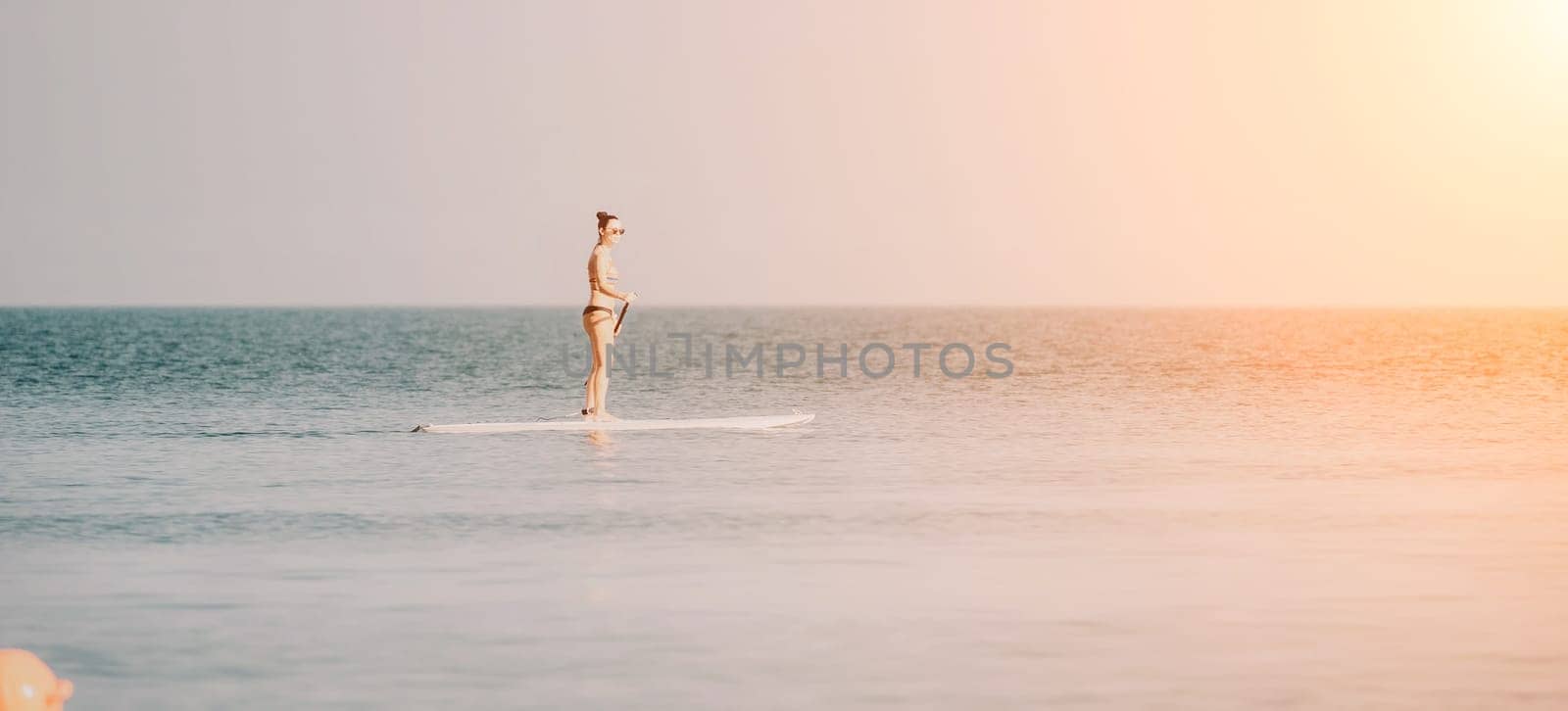 Sea woman sup. Silhouette of happy middle aged tanned woman in rainbow bikini, surfing on SUP board, confident paddling through water surface. Idyllic sunset. Active lifestyle at sea or river. by panophotograph