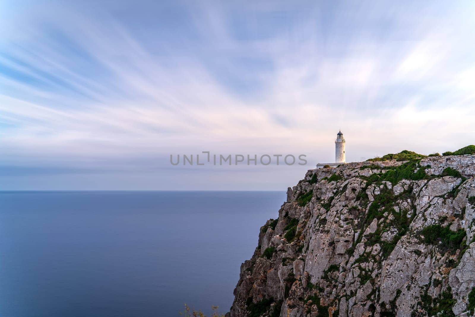 Long-exposure shot of a lighthouse on a cliff, featuring a smooth sea and fluffy clouds, conveys solitude.