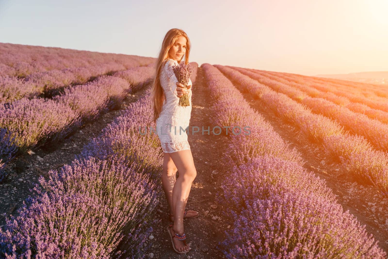 Close up portrait of young beautiful woman in a white dress and a hat is walking in the lavender field and smelling lavender bouquet.