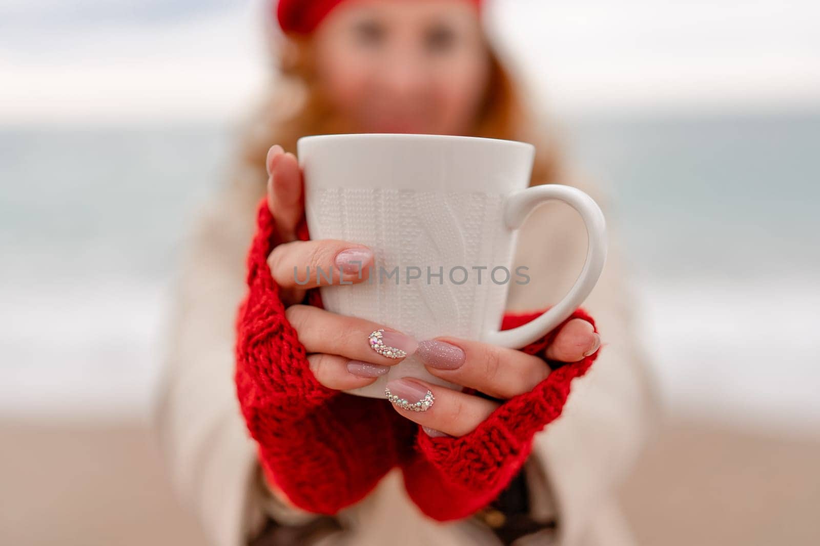 Woman beach in red beret scarf and mitts holding a cup of tea in his hands. Depicting beach relaxation and cozy attire. Walks by the sea.