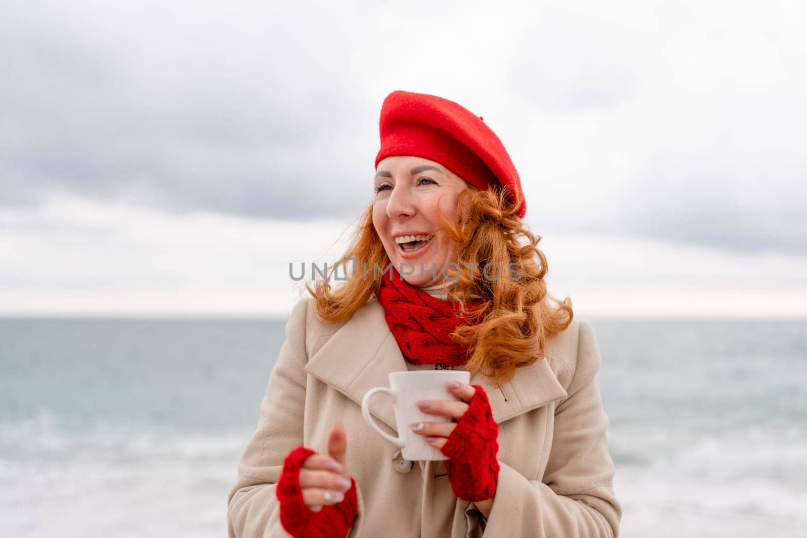 Woman beach in red beret scarf and mitts holding a cup of tea in his hands. Depicting beach relaxation and cozy attire. Walks by the sea by Matiunina