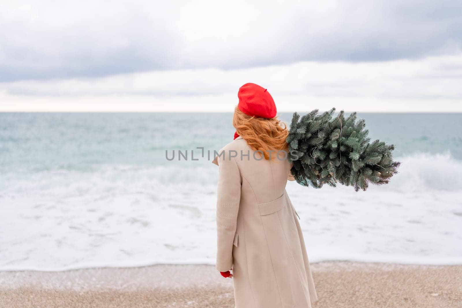 Redhead woman Christmas tree sea. Christmas portrait of a happy redhead woman walking along the beach and holding a Christmas tree on her shoulder. She is dressed in a light coat and a red beret. by Matiunina