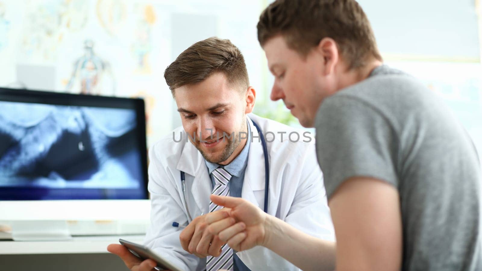 Portrait of smiling doc wearing white uniform with stethoscope. Patient looking to laptop attentively. Medical treatment and health care concept. Blurred background
