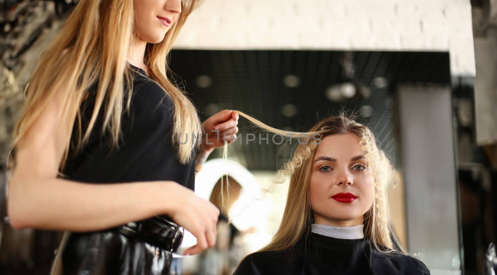 Beautiful Woman Getting Curly Hairstyle in Salon. Hairdresser Curling Hair for Young Female Client. Hairstylist Making Wavy Hairdo. Styling Curls for Girl with Red Lipstick Looking at Camera Shot