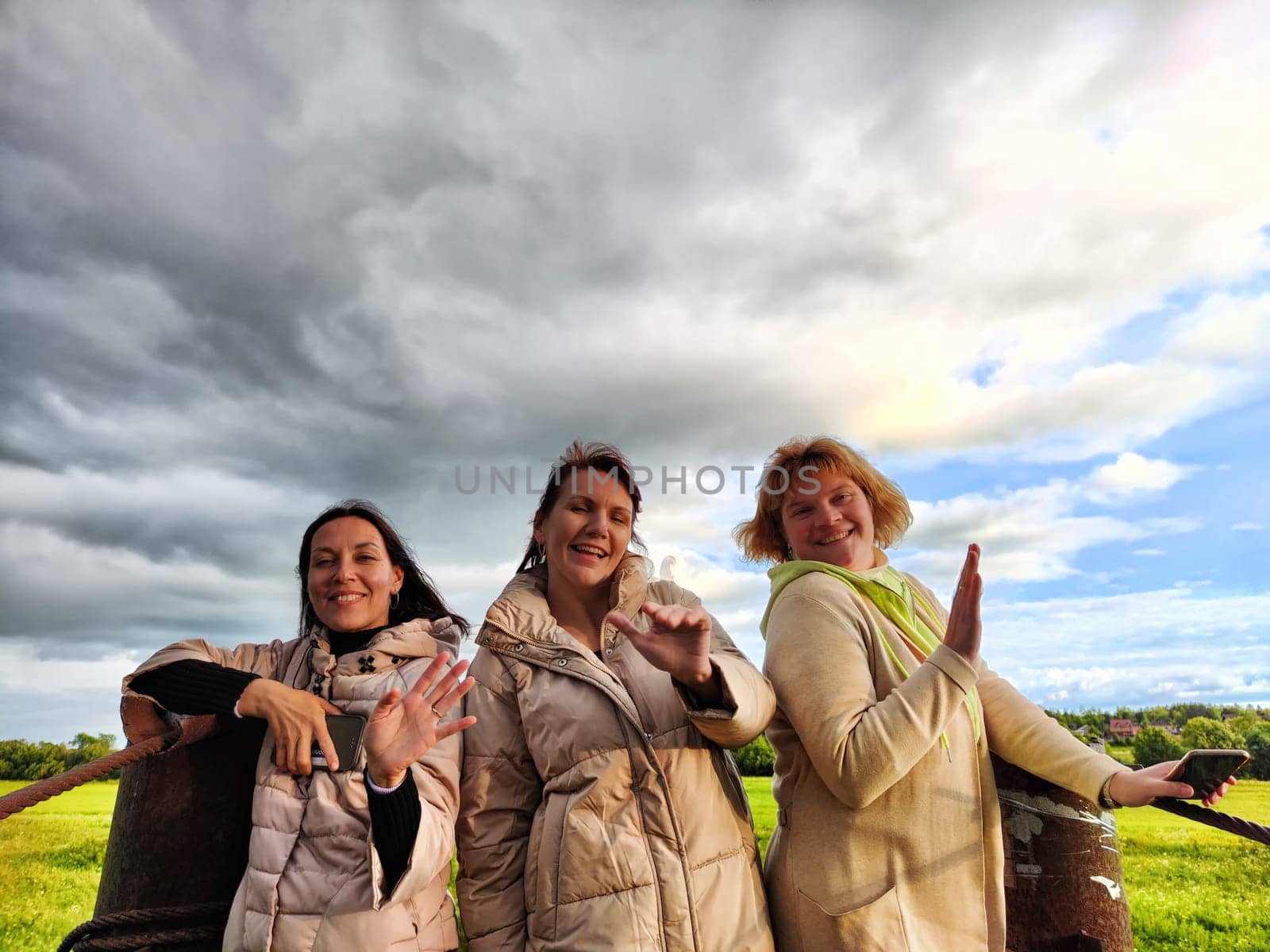 Three funny tourist girls on old bridge take selfies against the background of an alarming dramatic sky with thunderclouds. Middle-aged women having fun outdoors