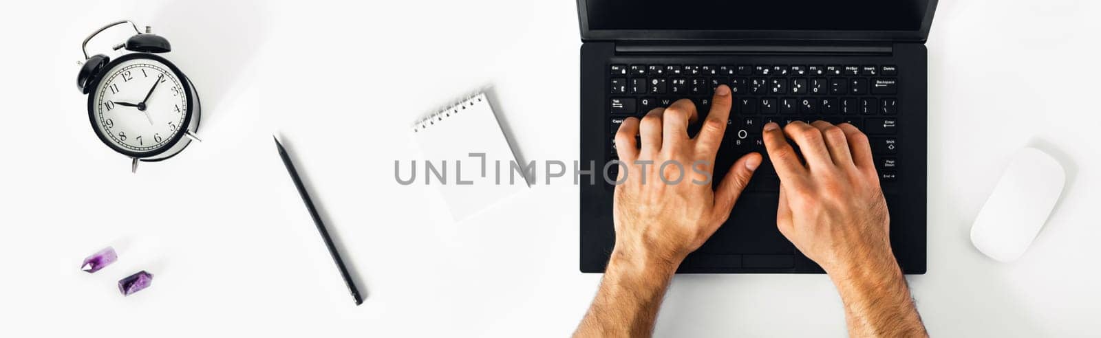 The man working at home on the remote. A black-and-white workplace with a laptop, alarm clock, notebook and men's hands.