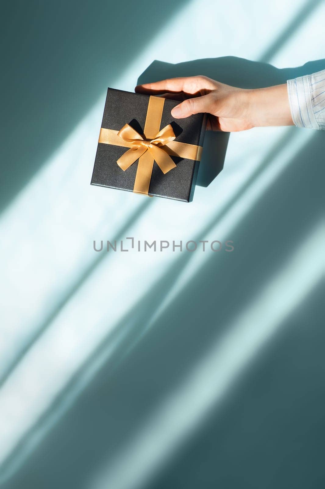 Vertical photo with gift box in a female hand on sunlights on blue background. Flat lay, top view, place for text.