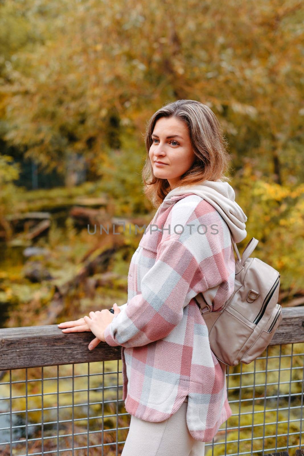 Portrait Of Cute Young Woman In Casual Wear In Autumn, Standing On Bridge Against Background Of An Autumn Park And River. Pretty Female Walking In Park In Golden Fall. Copy Space. Smiling Girl In The Park Standing On Wooden Bridge And Looking At The Camera In Autumn Season by Andrii_Ko