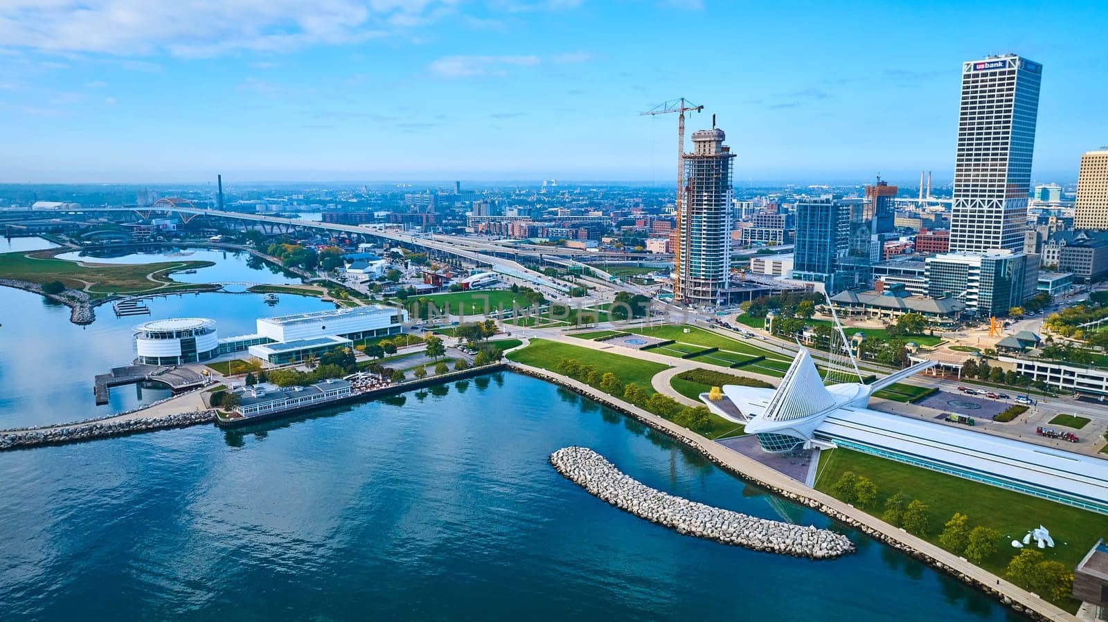 Aerial view of Milwaukee's vibrant waterfront cityscape featuring a modern suspension bridge, the glassy Quadracci Pavilion and a rising skyscraper under construction, depicting urban growth and harmony with nature.