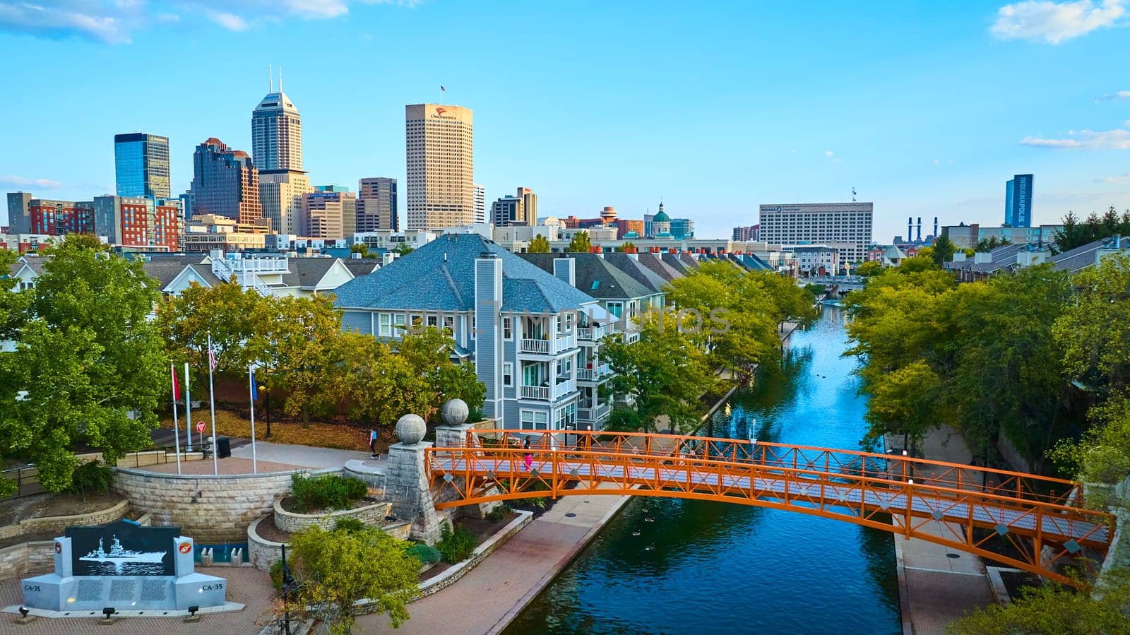 Vibrant 2023 Indianapolis cityscape showcasing a blend of urban development and residential charm, captured during golden hour from a drone perspective, highlighting the orange pedestrian bridge, serene canal, and dynamic skyline.