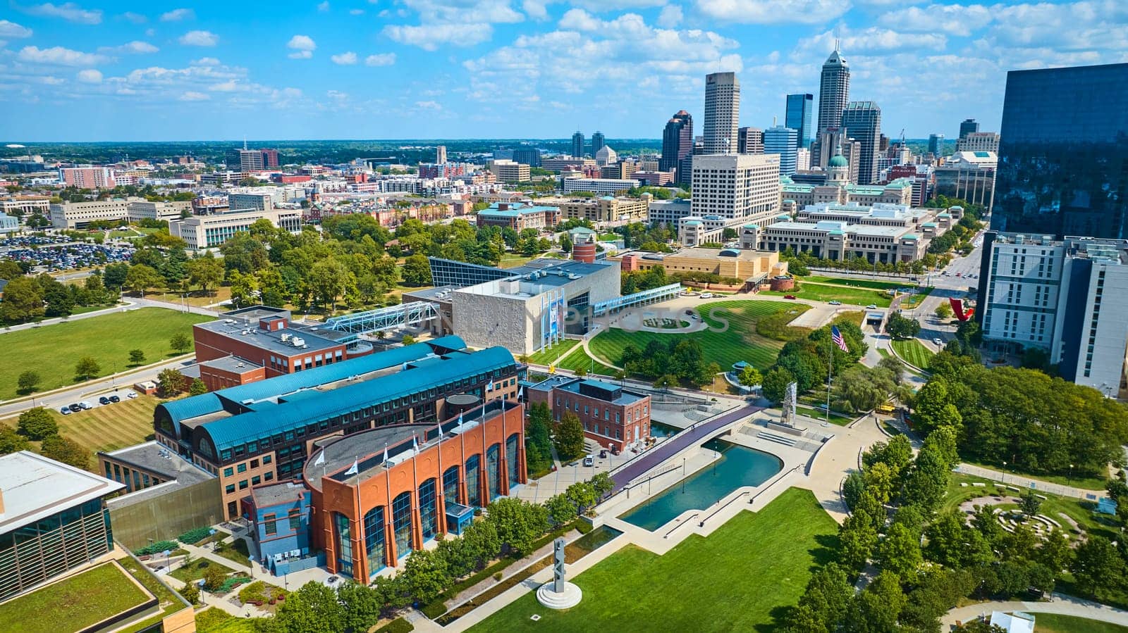 Vibrant aerial view of modern Indianapolis cityscape, featuring diverse architectural styles, green urban park, and a bustling river canal captured by a drone in 2023