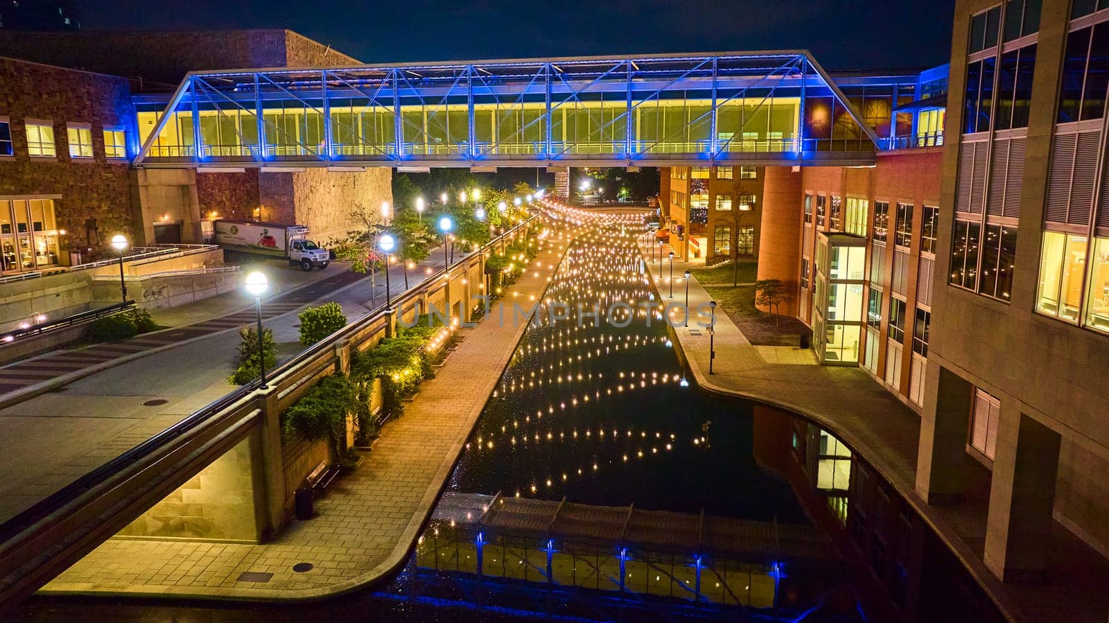 Nighttime aerial view of a vibrant Indianapolis cityscape, showcasing a lit pedestrian bridge crossing the canal, surrounded by modern architecture.