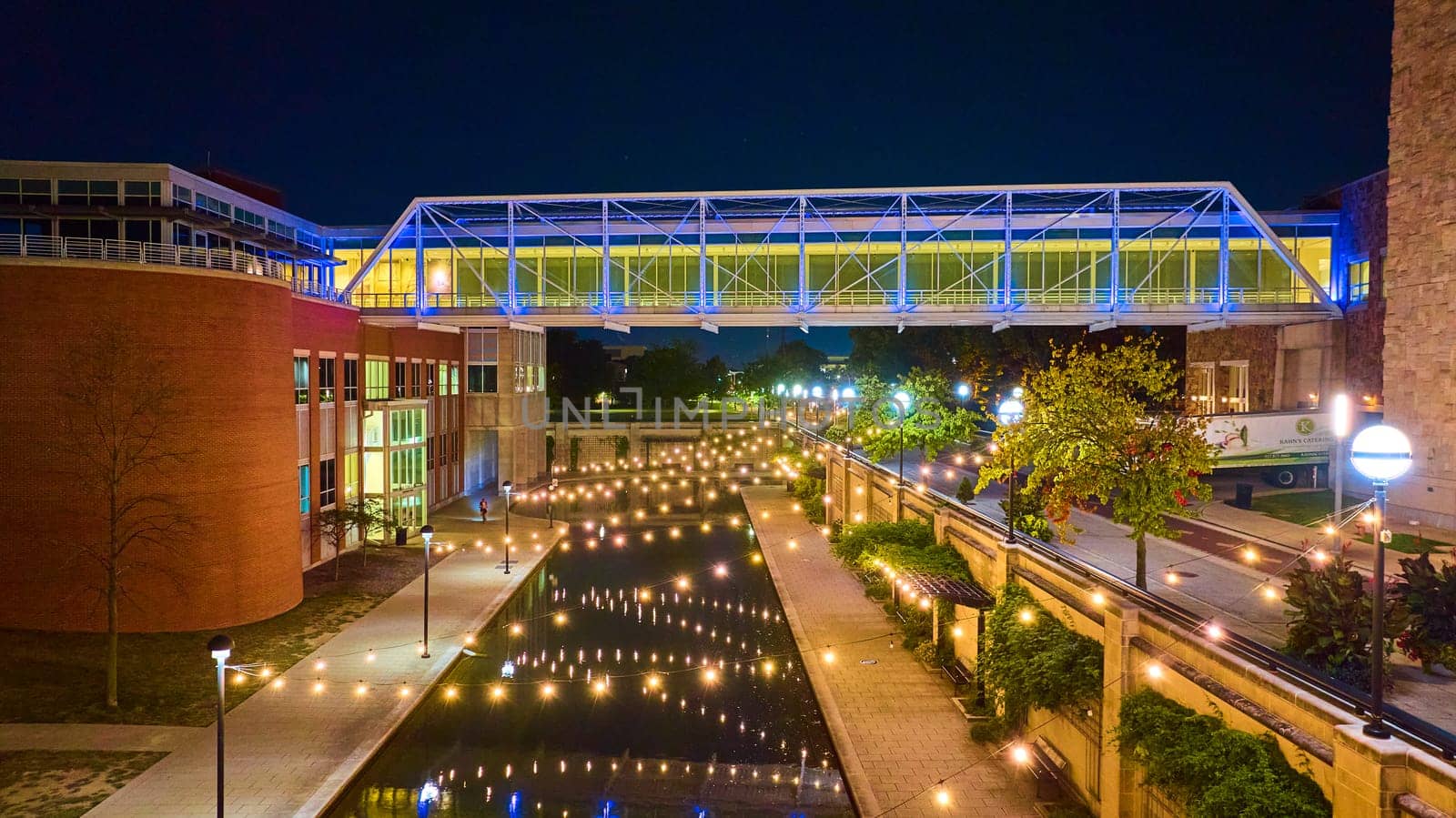 Evening aerial shot of modern illuminated pedestrian bridge, reflective canal, and vibrant campus walkway in Indianapolis, 2023