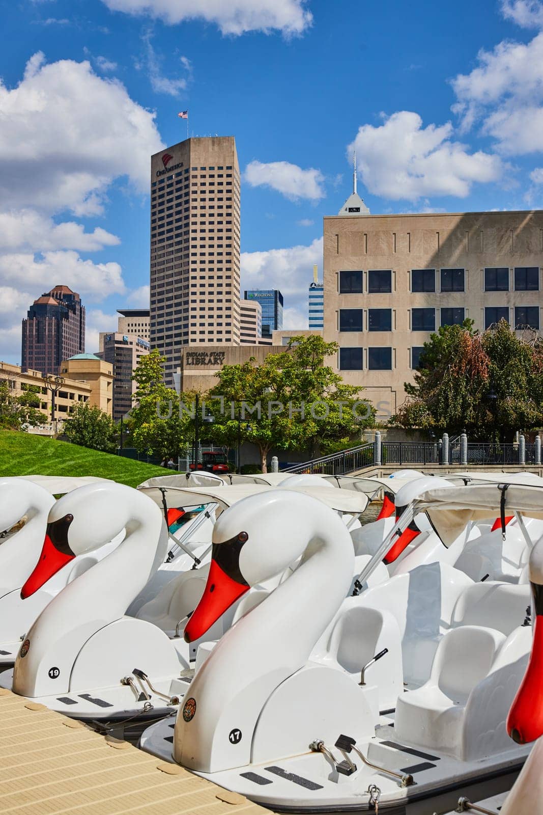 Sunny day in Indianapolis with colorful swan pedal boats awaiting riders on the canal, contrasted against the urban skyline, 2023