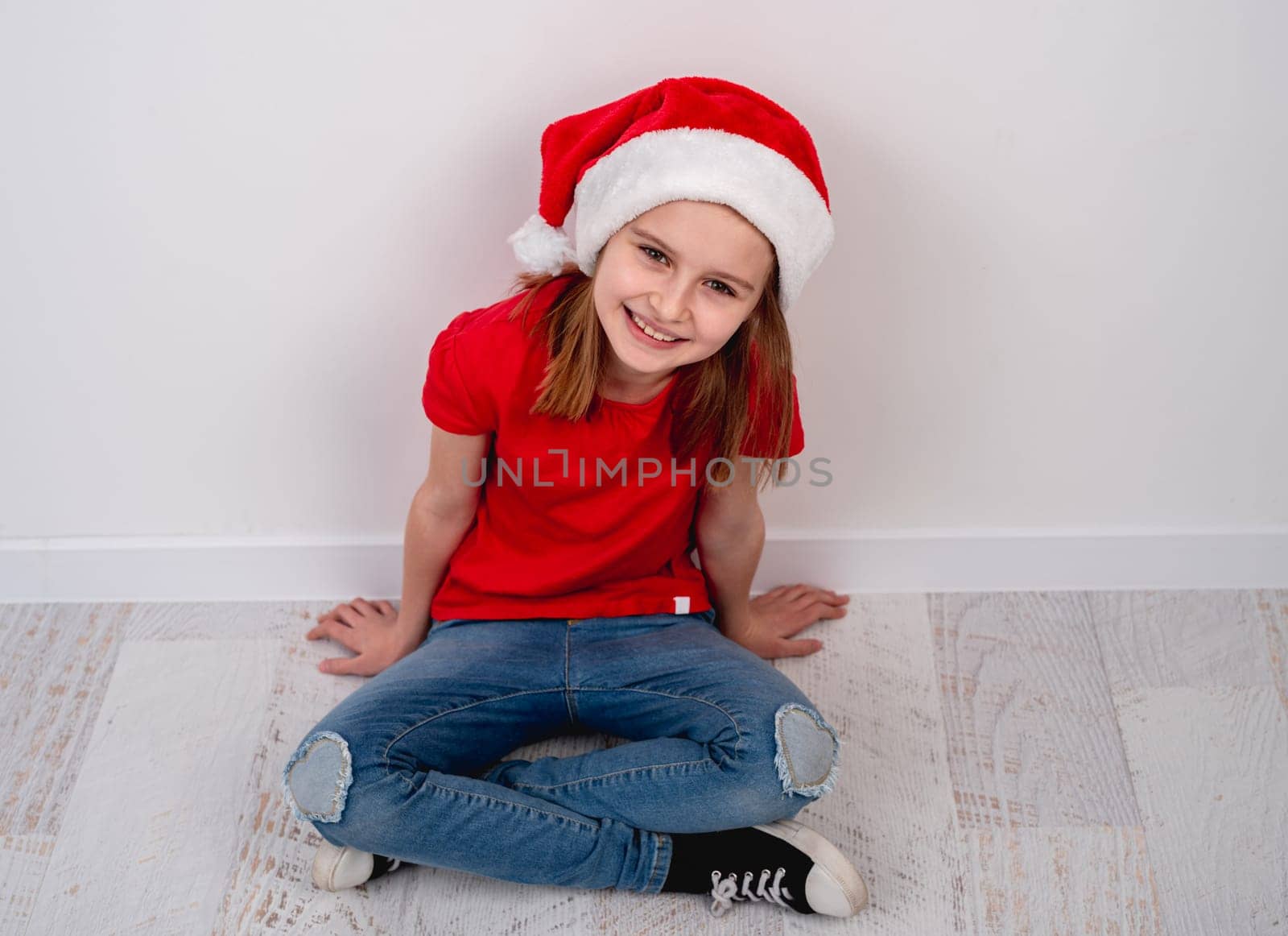 Little Girl In Red Shirt And Santa Hat Sitting On Floor For Christmas Photo On White Background by tan4ikk1