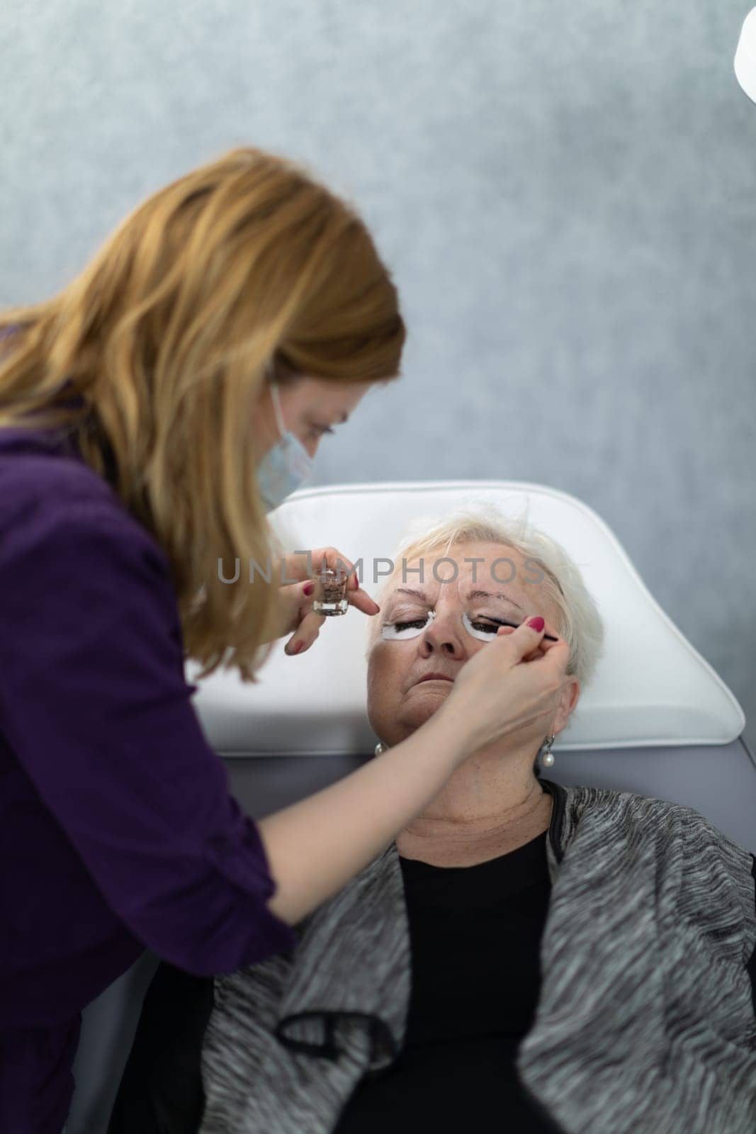 A young woman works as a cosmetologist at a beauty salon. She applies henna to her client's eyelashes.