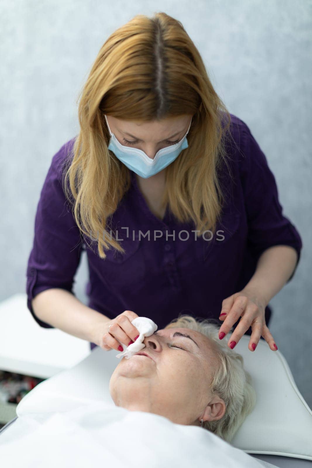 A patient of an aesthetic clinic is lying on a bed and talking to a cosmetologist standing next to her.