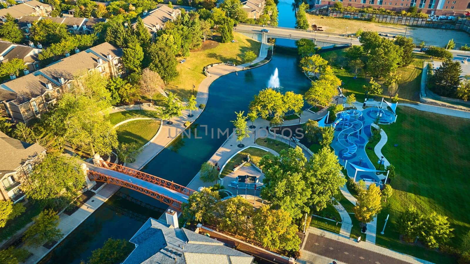 Aerial View of Bustling Suburban Park at Golden Hour in Indianapolis, Featuring River, Playground and Pedestrian Bridge