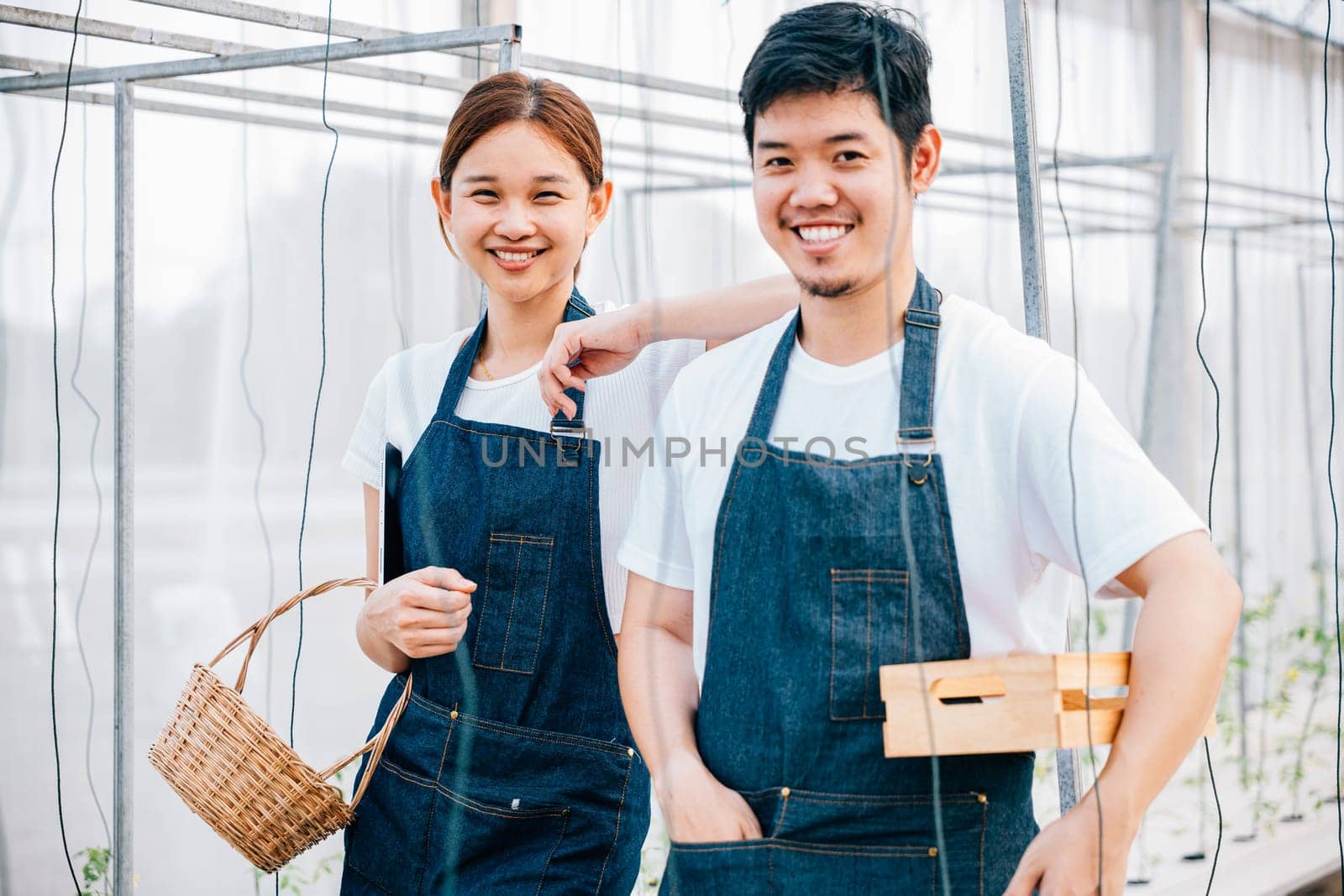 In a greenhouse an Asian woman and man a smiling farming couple hold fresh tomatoes and vegetables. Their portrait reflects joy and confidence in their occupation emphasizing nature bountiful growth.