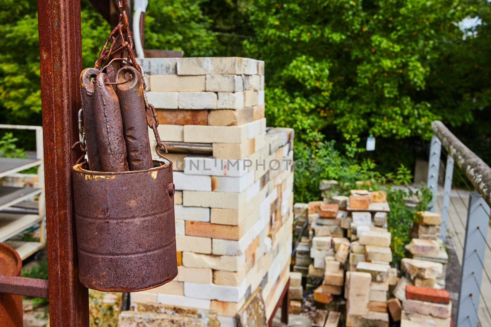 Rusted metal bucket at a sunny construction site in Indianapolis, featuring a stack of bricks and green foliage