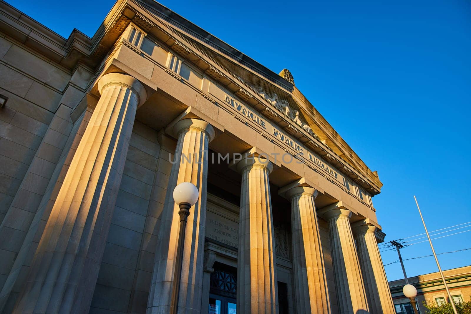 Golden hour sunrise illuminates the grand architecture of Muncie Public Library, Indiana, showcasing its ornate columns and stonework against a clear blue sky.