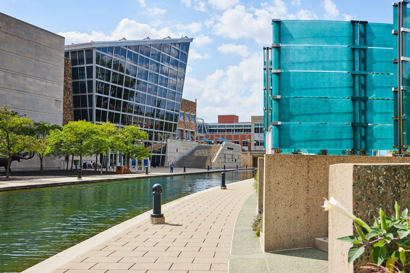 Sunny day in Indianapolis showcasing modern architecture, a serene canal and vibrant teal structure alongside a peaceful waterside walkway.