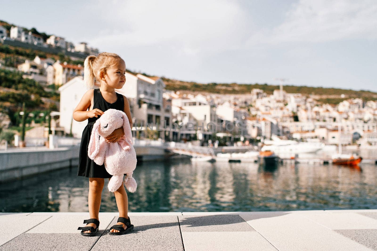 Little girl with a big pink plush hare stands on the pier against the background of the marina with yachts. High quality photo