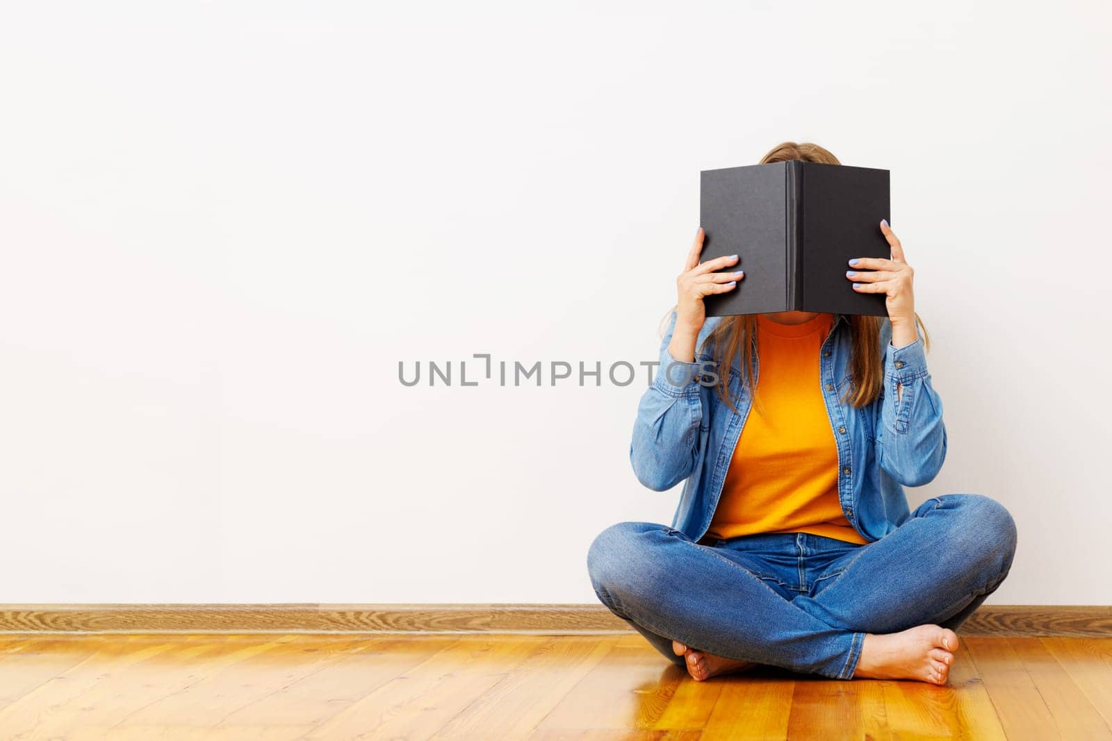 A young woman covers her face with a book as she sits on floor against the wall by andreyz