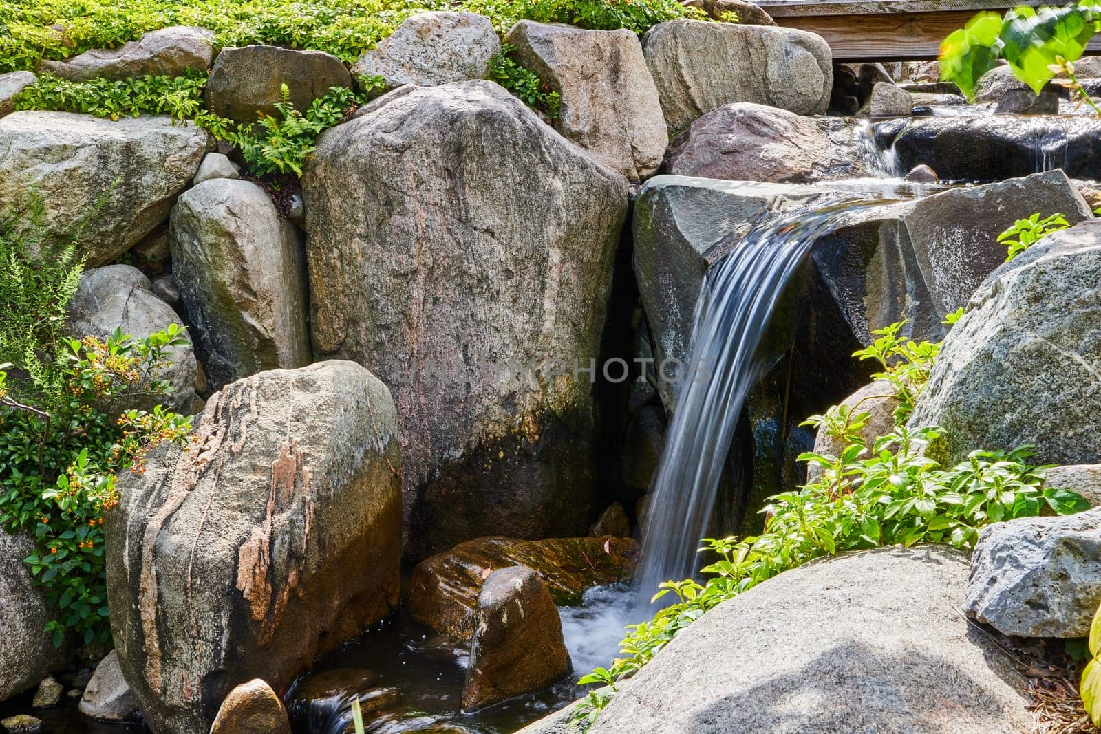 Peaceful man-made waterfall streaming over rugged boulders amidst lush greenery, a tranquil oasis in Elkhart's Botanic Gardens, Indiana, 2023.