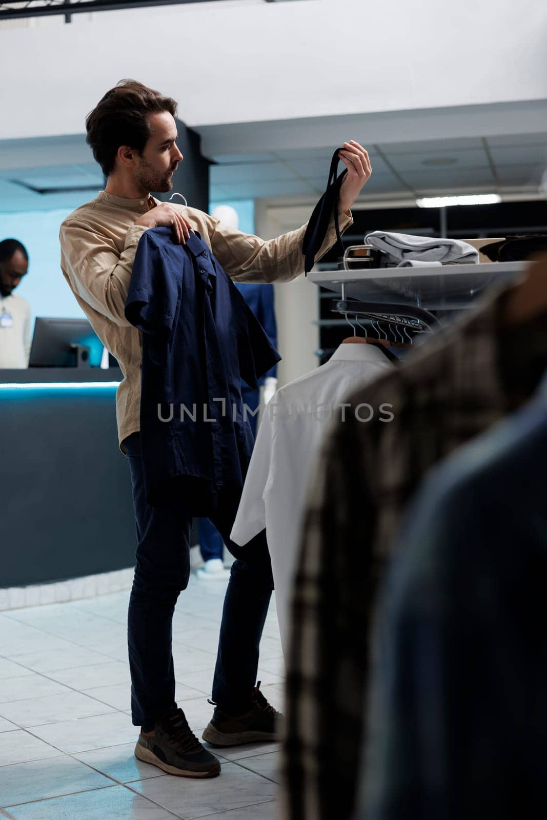 Client examining tie and shirt style while choosing formal outfit in shopping mal menswear department. Young caucasian man holding apparel and accessory in fashion clothing store