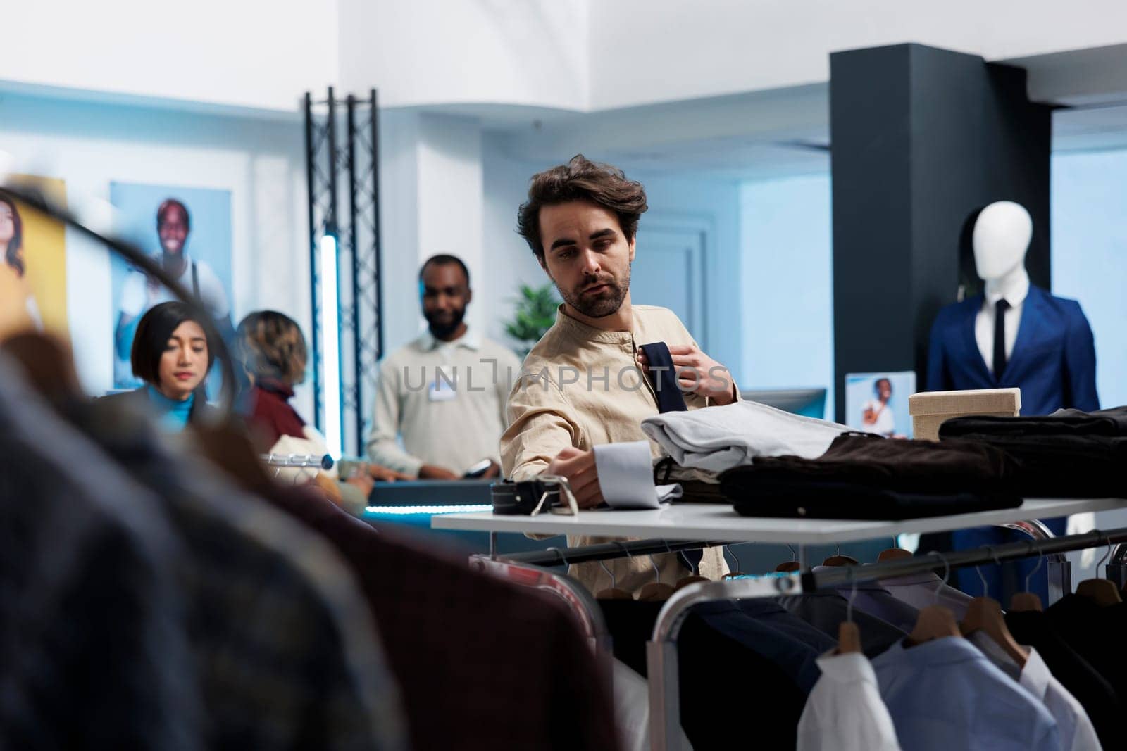 Young man choosing stylish tie and examining options while shopping in clothing store. Fashion boutique customer selecting formal trendy accessory in menswear retail department