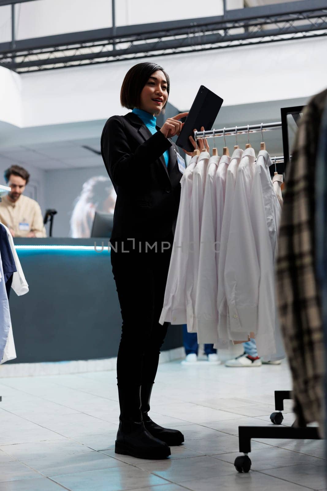 Shopping mall employee managing shirts new collection inventory using digital tablet. Young asian woman checking apparel stock showcasing on display rack while working in clothing store