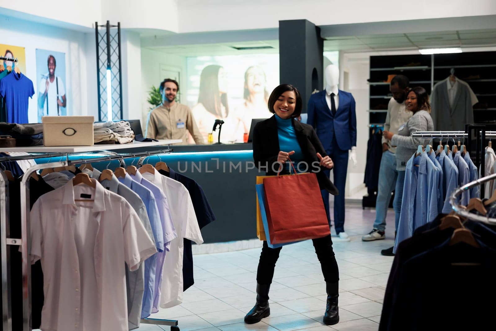 Smiling asian woman dancing with purchases, enjoying buying apparel in clothing store. Boutique cheerful customer holding packages and making moves to music in shopping center