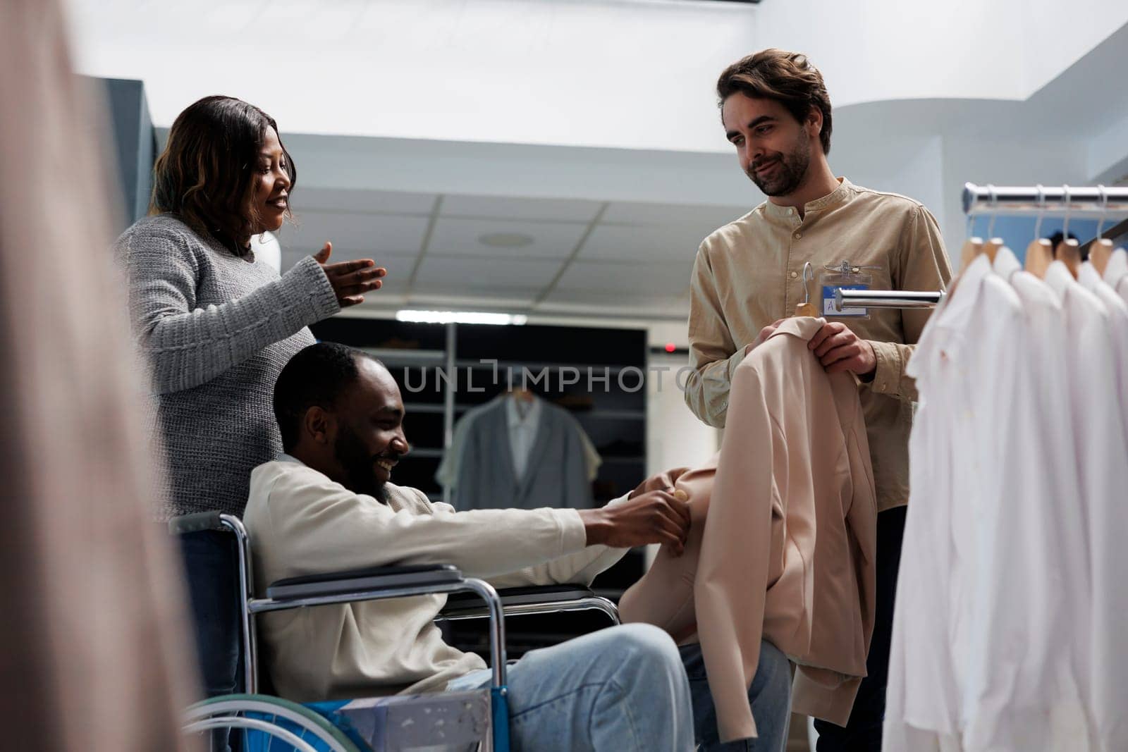 African american man in wheelchair shopping for womenswear with wife, getting help from clothing store employee. Mall customer with physical disability choosing jacket for girlfriend