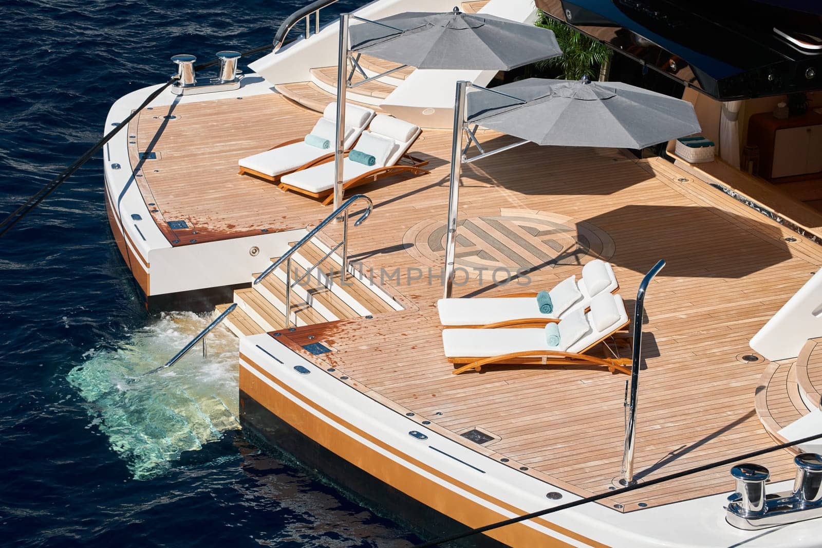 Close-up view of a relaxation area on the open teak deck on the stern of an expensive huge mega yacht at sunny day, stairs to water, wealth life, lounge chairs by vladimirdrozdin