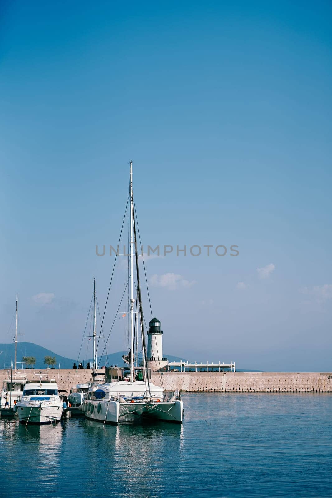 Yachts and a sailing catamaran moored near a breakwater with a lighthouse. Lustica Bay, Montenegro by Nadtochiy