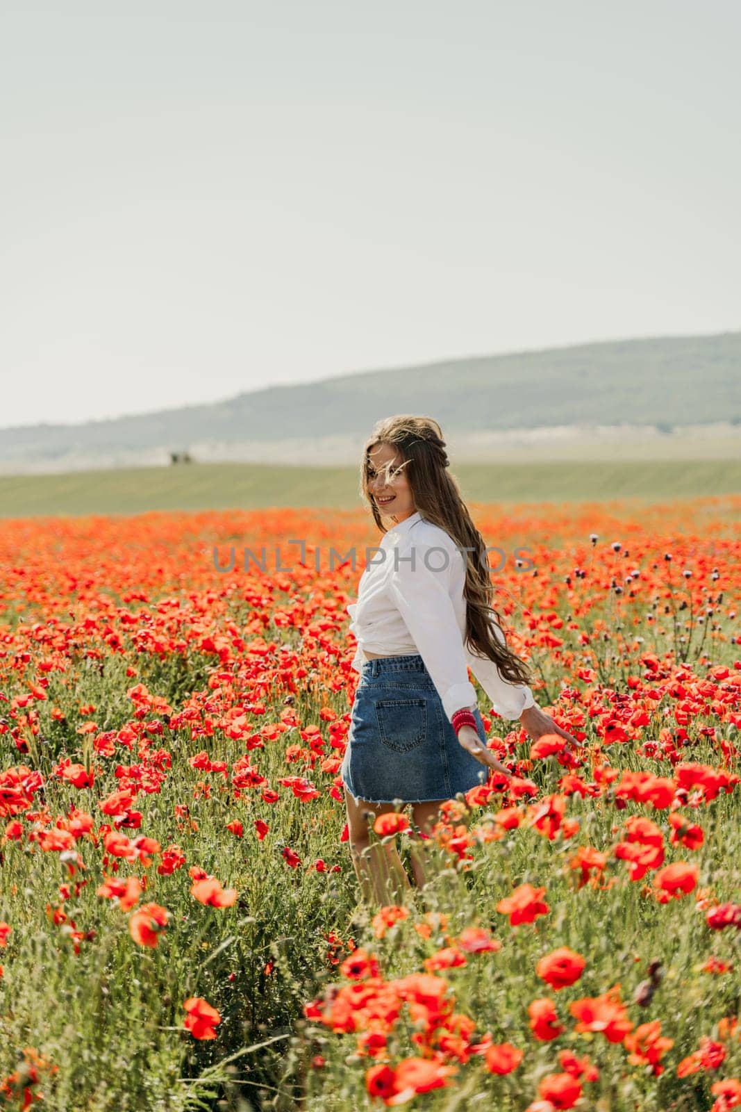 Happy woman in a poppy field in a white shirt and denim skirt with a wreath of poppies on her head posing and enjoying the poppy field. by Matiunina